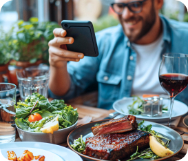 A man taking a photo of a meal with steak, salad, and a glass of red wine on a table.