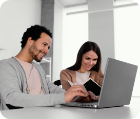 Two professionals working on a laptop in an office setting. Male in grey cardigan and pink shirt, female in beige cardigan and white top