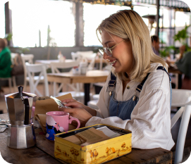 A woman sitting at a table in a restaurant, holding her phone with a pink mug on the table.